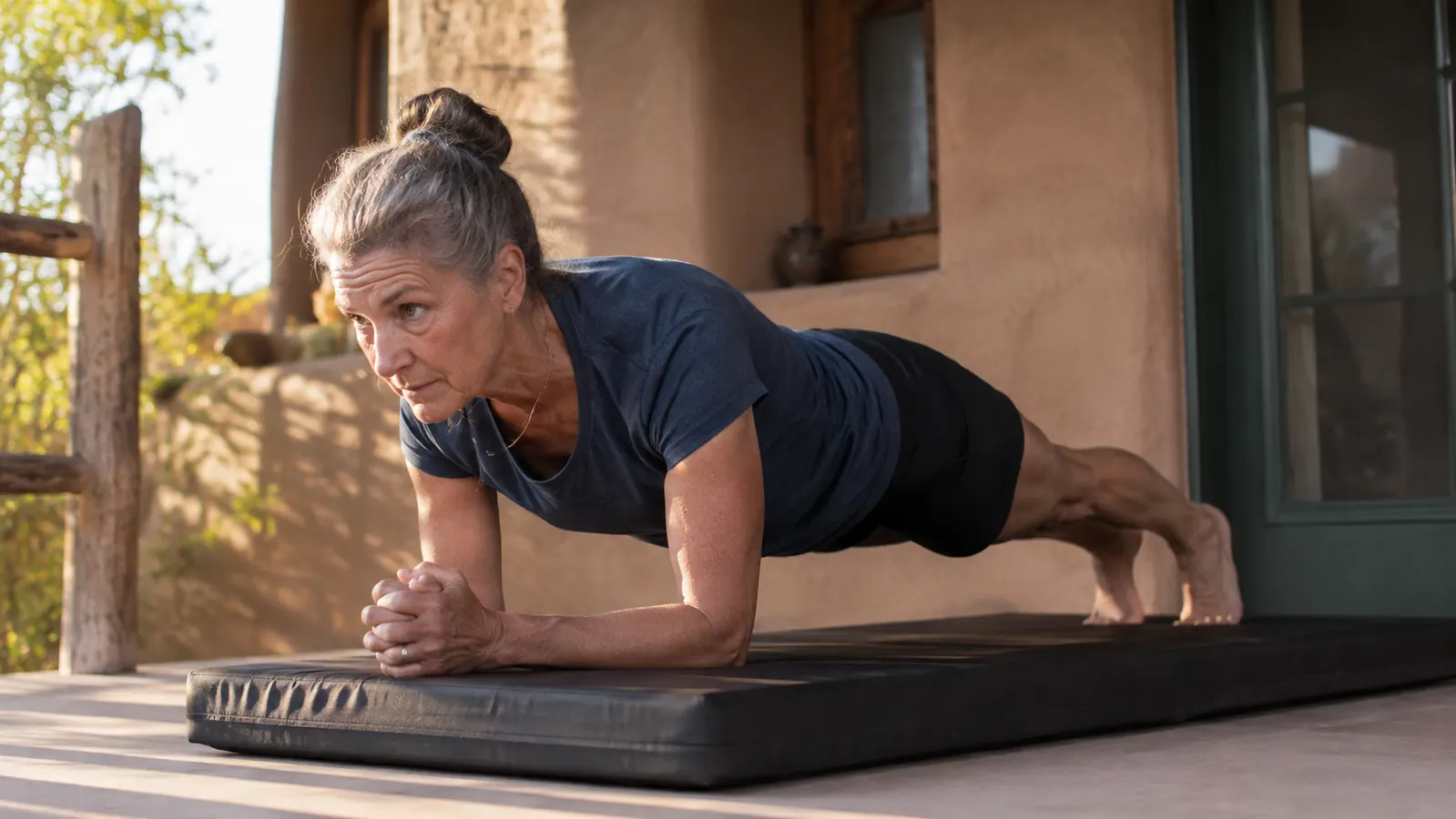 A woman in her fifties kneeling on a soft mat on a porch, palms flat on the floor in front of her, early morning light over a desert yard